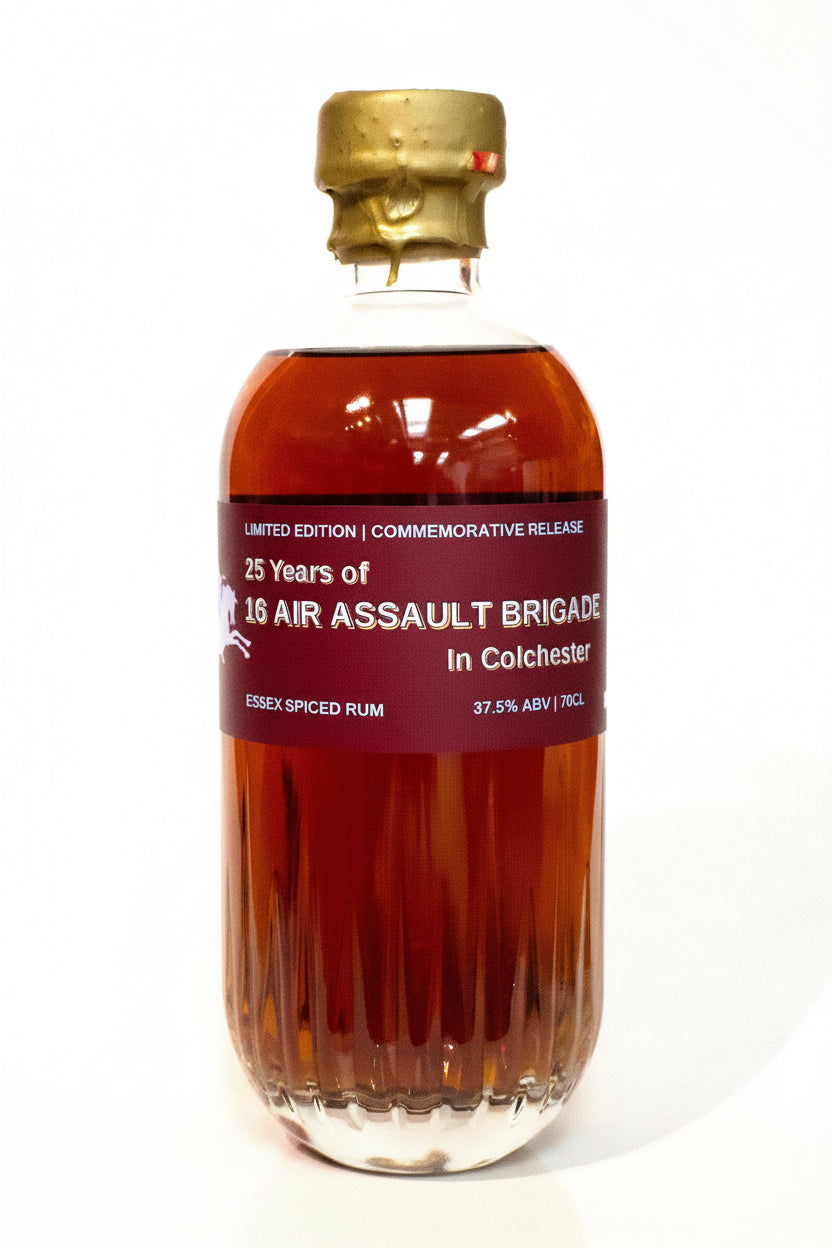 A bottle of commemorative Essex Spiced Rum with amber liquid, a gold cap, and a dark red label reading '25 Years of 10th Air Assault Brigade in Colchester'.