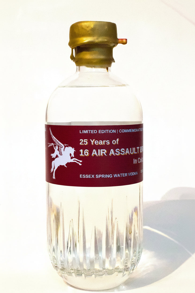 A clear glass bottle of Essex Spring Water Vodka with a gold cap and a dark red label featuring a white winged horse logo and text celebrating 25 years of 16 Air Assault 50ml shots.