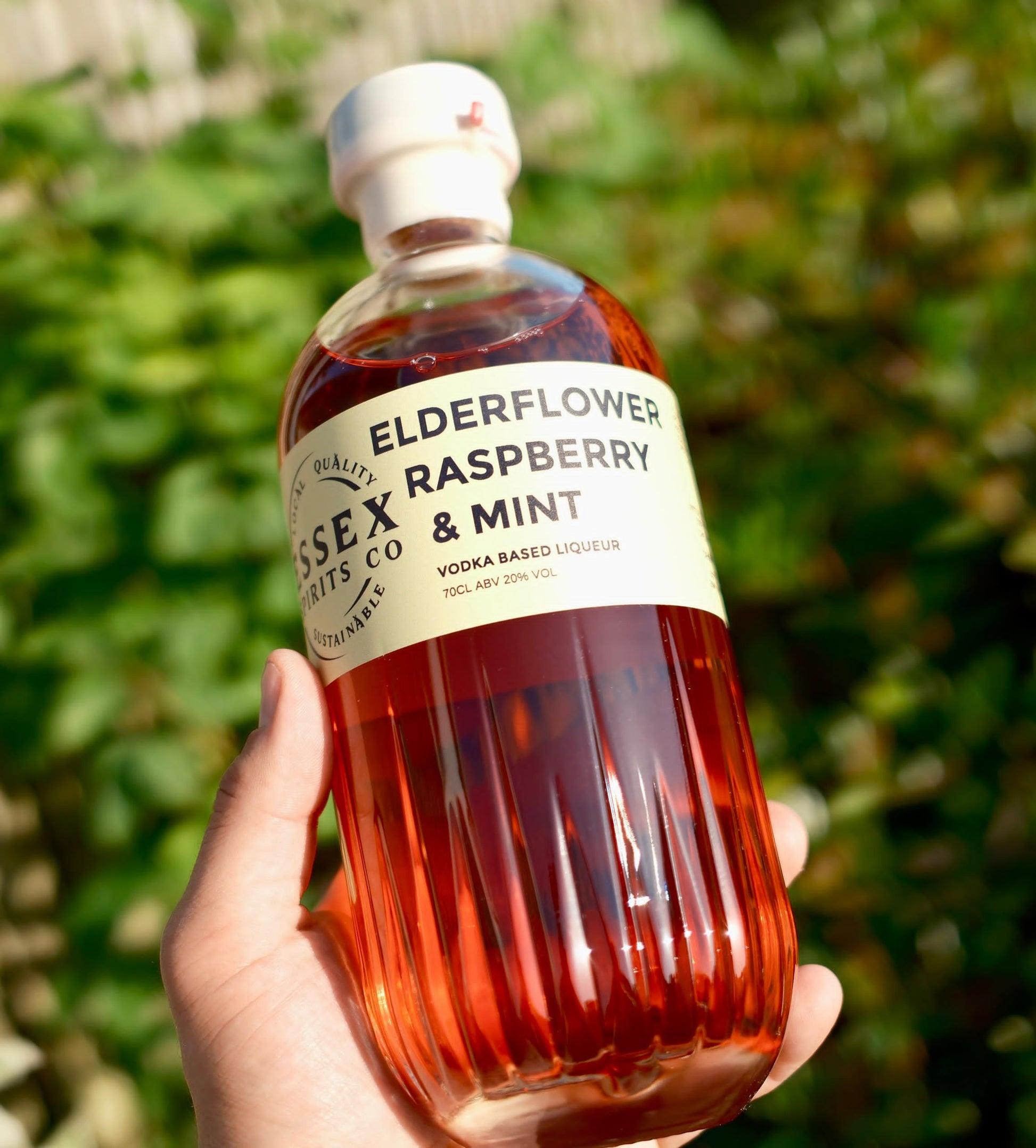 Bottle of Essex Spirits Co. Elderflower Raspberry & Mint liquor held in a hand with a blurred green background